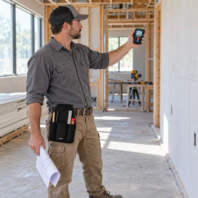 Construction professional measuring an interior wall with a Makita laser distance measure while wearing an Agoz utility tool pouch on his waist belt.